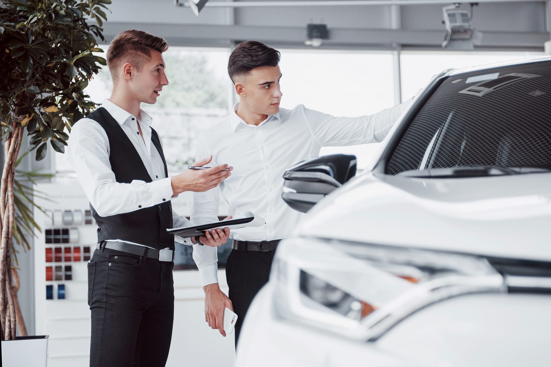 Two men stand in the showroom against cars. Close-up of a sales manager in a suit that sells a car to a customer. The seller gives the key to the customer.